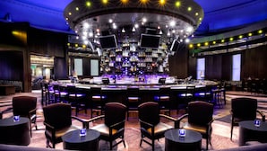 Un espace bar doté d’un comptoir circulaire, de plusieurs écrans et d’une grande variété de bouteilles. On y trouve également plusieurs chaises et tables disposées autour du bar.