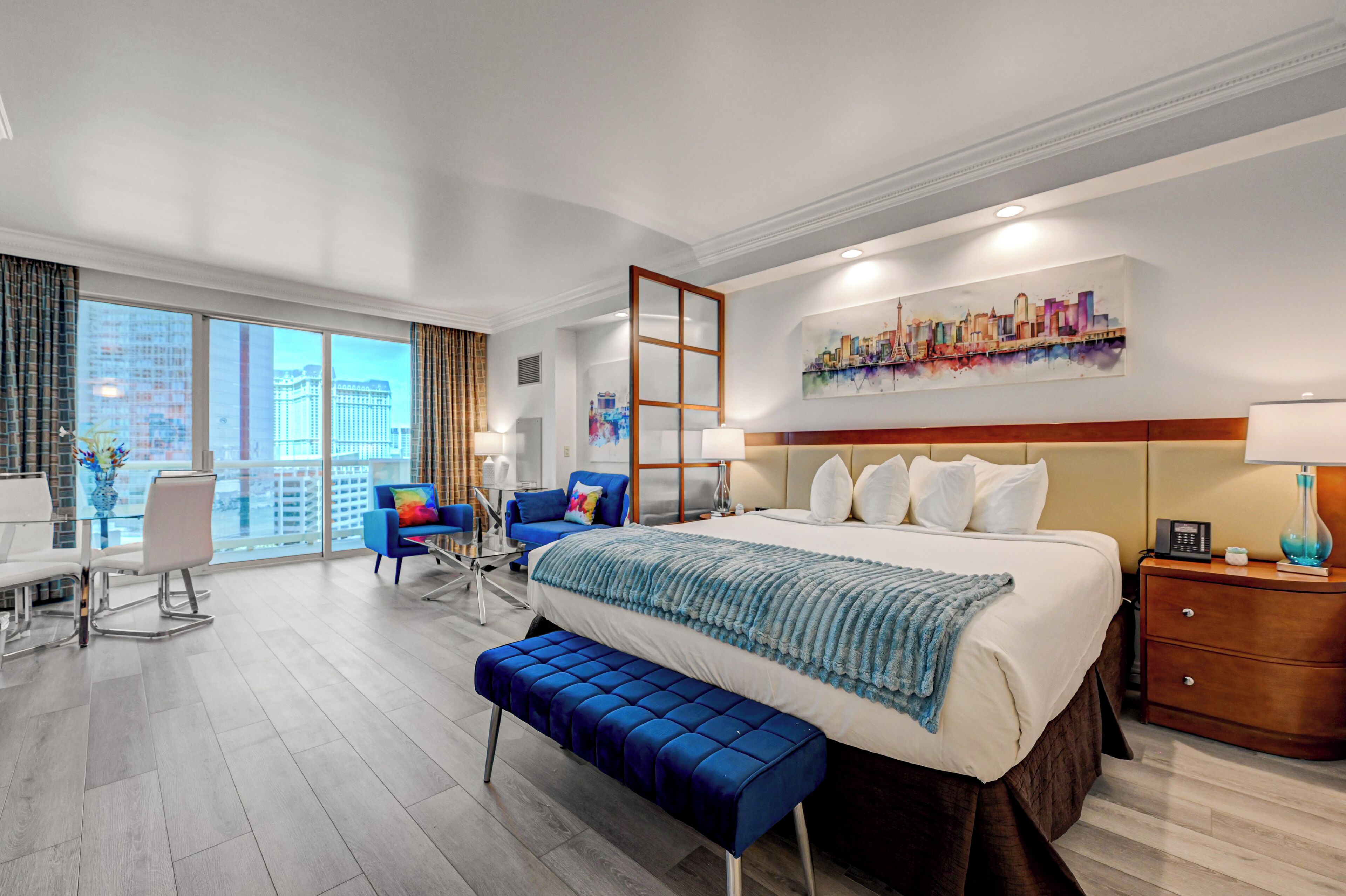 A hotel room with a large bed, a desk, a chair, a bench, and a cityscape view.
