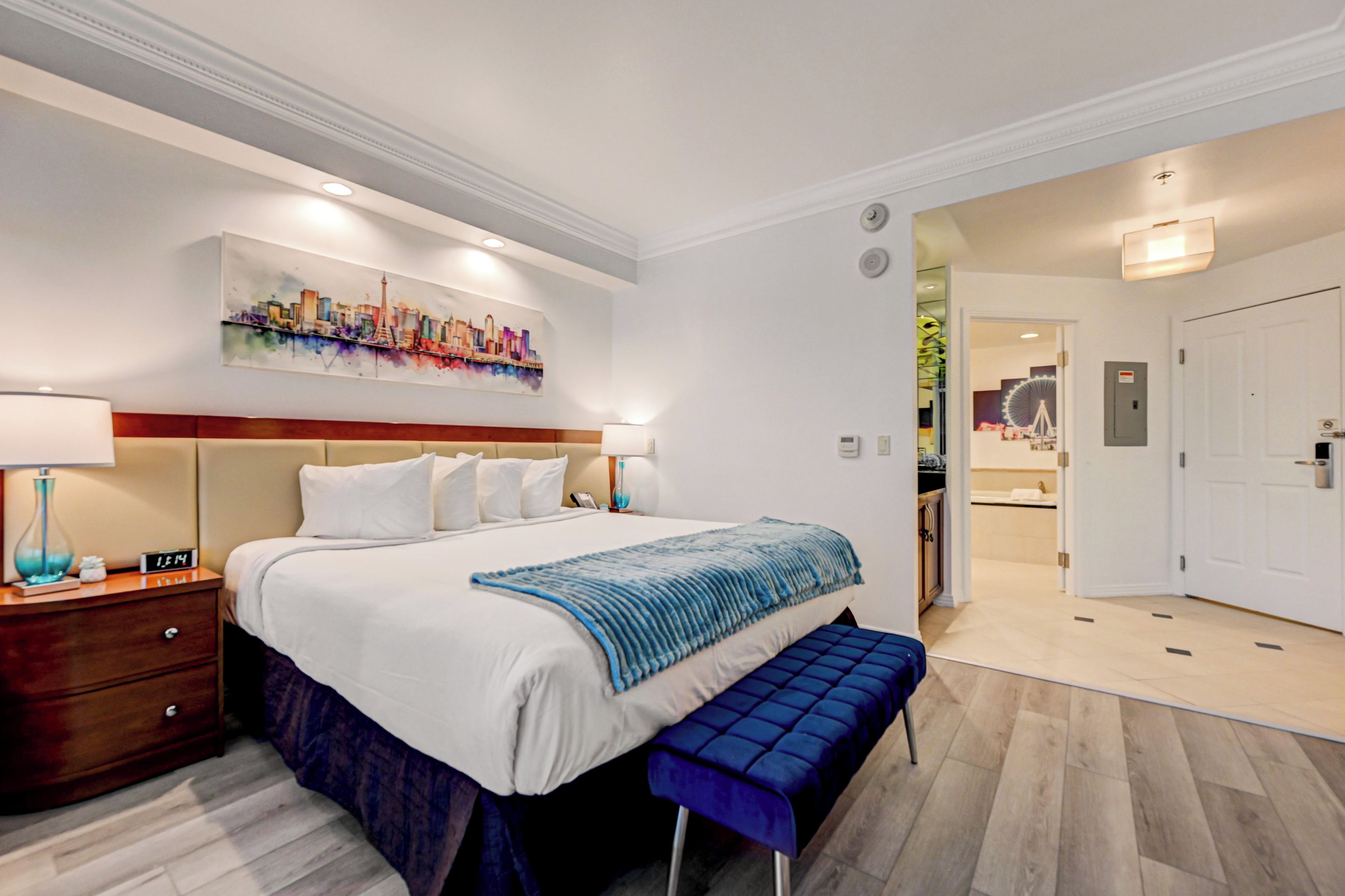 Quarto de hotel com cama, mesas de cabeceira, um banco, um quadro e banheiro visível por uma porta aberta.