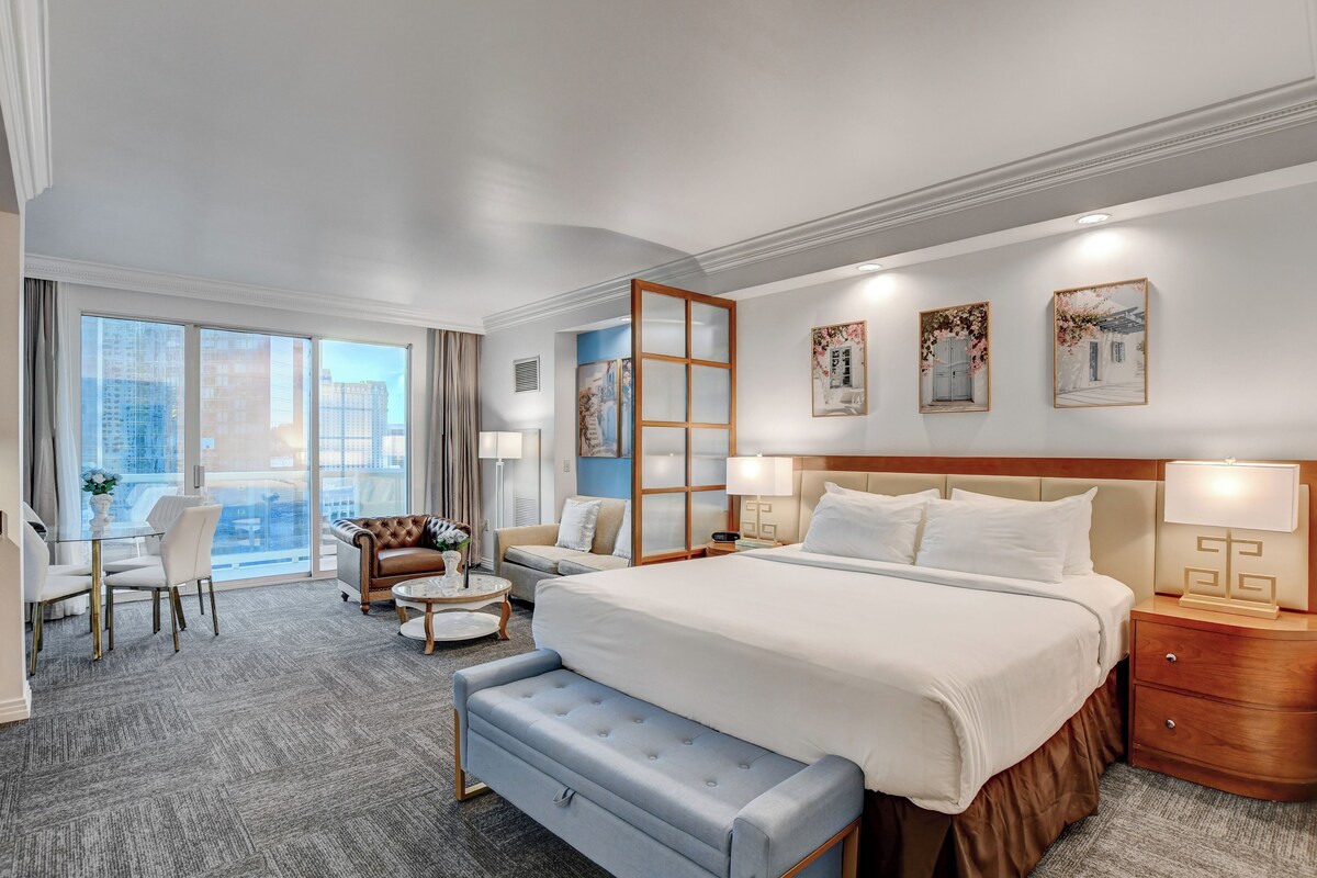 Een hotelkamer met een groot bed, een zithoek met een bank en fauteuils, een bijzettafeltje en uitzicht op de stad door grote ramen.