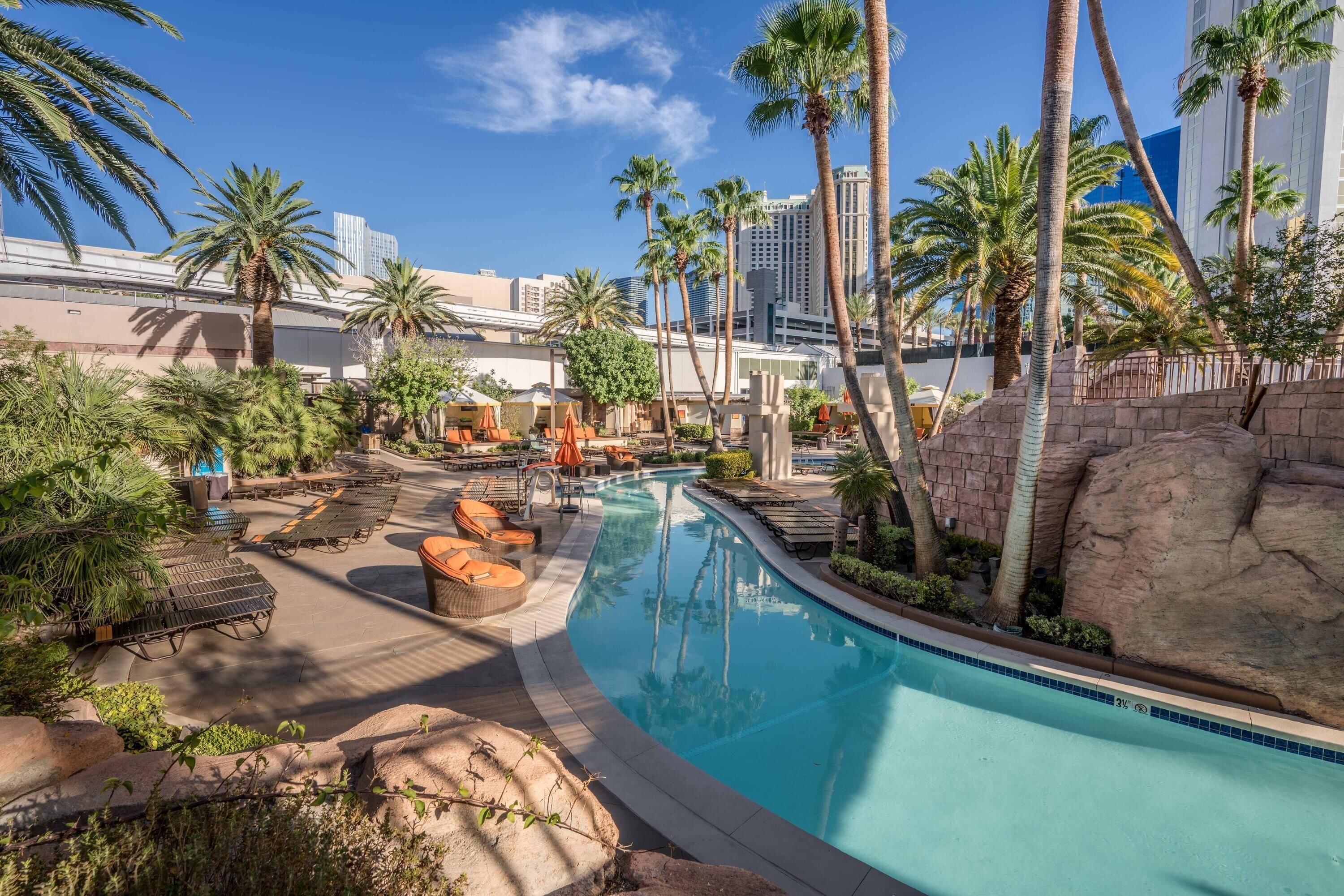 A pool area with lounge chairs, palm trees, and a view of a city skyline.