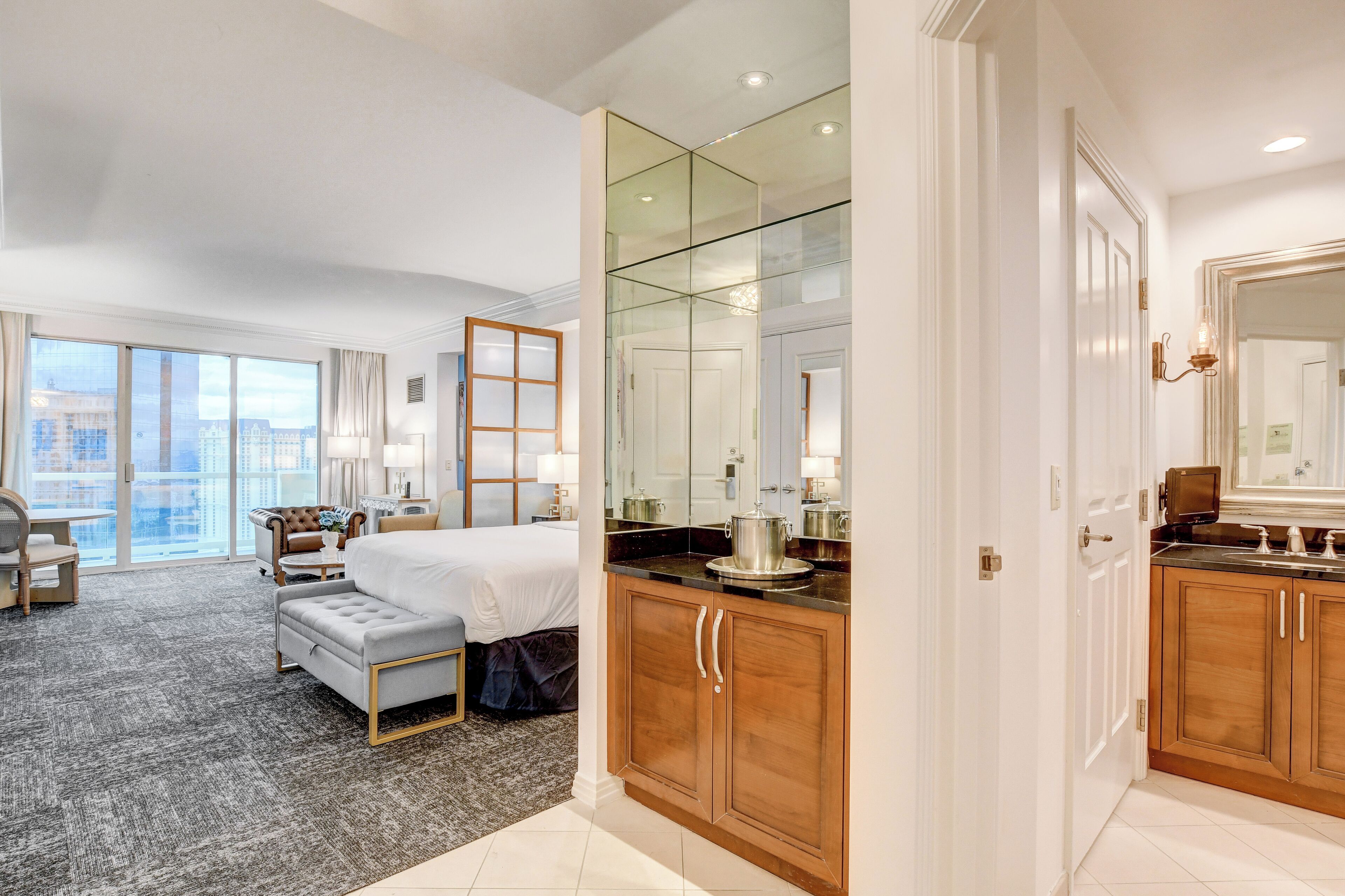 A hotel room with a bed, a seating area, a bathroom with a mirror, and a view of the city.
