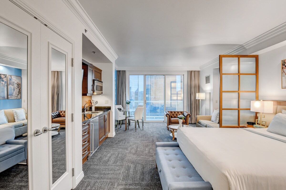 A hotel room with a large bed, a seating area with a sofa and chairs, a dining table, a kitchen area, and a balcony with a view.