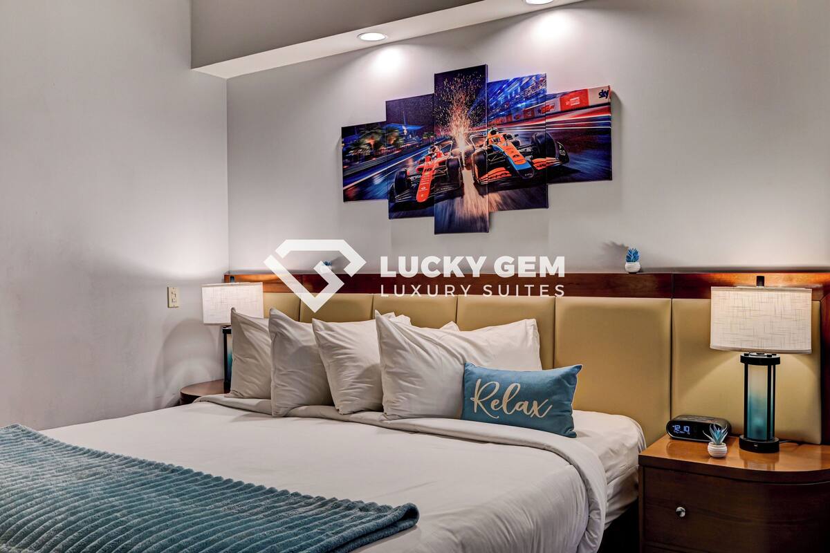 Een luxe suite met een bed, kussens, een nachtkastje met een lamp, een kunstwerk aan de muur, en een logo voor Lucky Gem Luxury Suites.