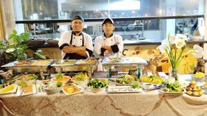 Uma mesa de buffet com diversos alimentos e dois chefs posicionados atrás dela.