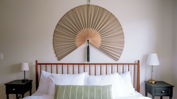 Een net opgemaakt bed met witte en groene kussens, een blauw beddegoed met patroon en een grote wandventilator.