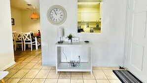 Ein helles, weißes Interieur mit einer Uhr, einem kleinen Tisch mit einer Pflanze und Blick in eine Küche durch ein Fenster.