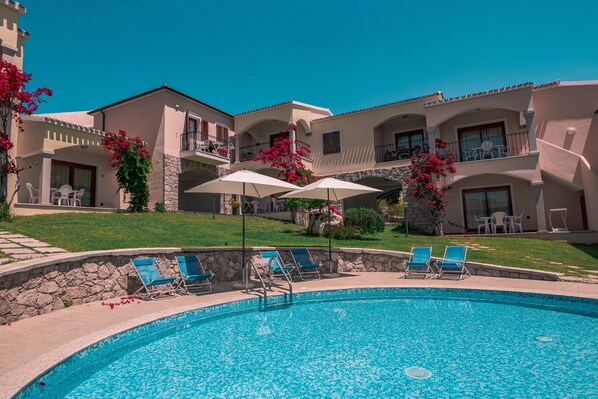 A pool area with lounge chairs and umbrellas, a stone wall, and a building with balconies.