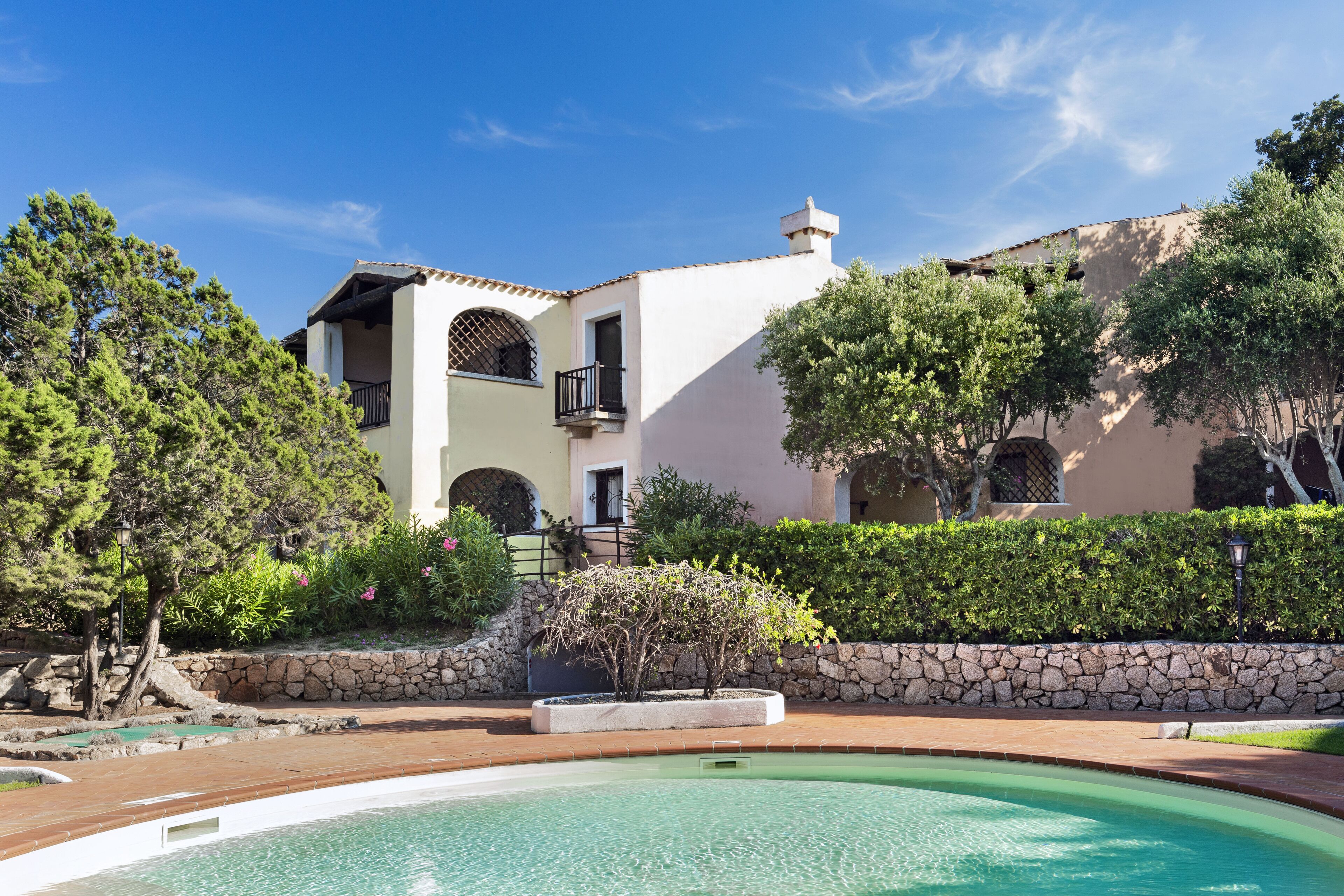 A two-story building with a pool, stone wall, and greenery.