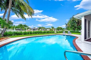A swimming pool with a clear blue water, surrounded by a white fence, palm trees, and a clear sky. - Waterfront, Hot Pool, Lounge Patio, Beach House (Palm Beach Gardens)