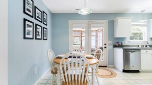 A dining area with a round table, white chairs, and a wooden floor. There are framed pictures on the wall, a ceiling light, and a kitchen area with a dishwasher and cabinets. - Charming Home! Just minutes From the Beach, Near Shopping, Dog Friendly (Oak Island)