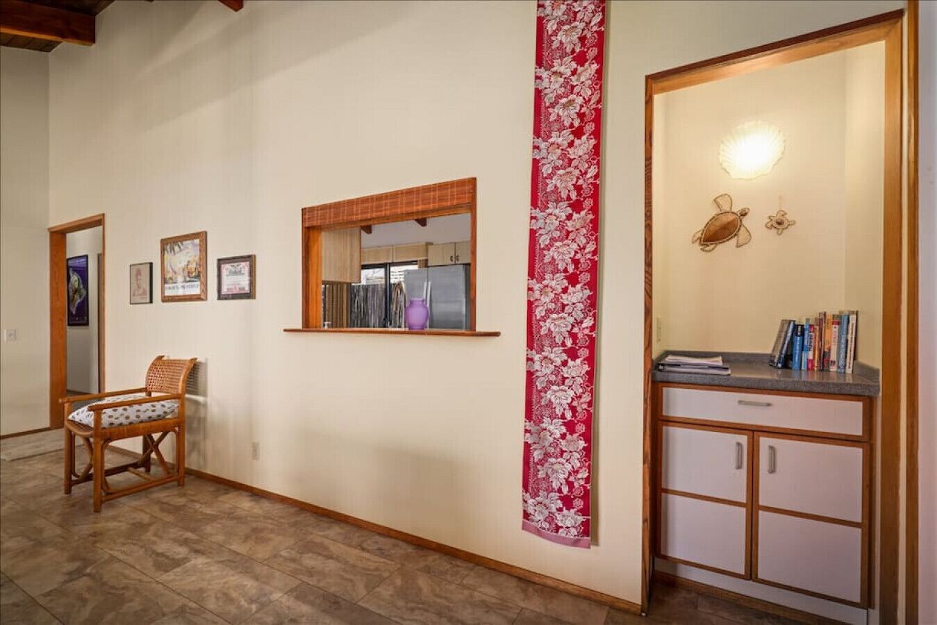 A hallway with a wooden chair, a mirror, framed pictures, a window, a red floral wall hanging, and a cabinet with books.