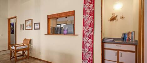 A hallway with a wooden chair, a mirror, framed pictures, a window, a red floral wall hanging, and a cabinet with books.
