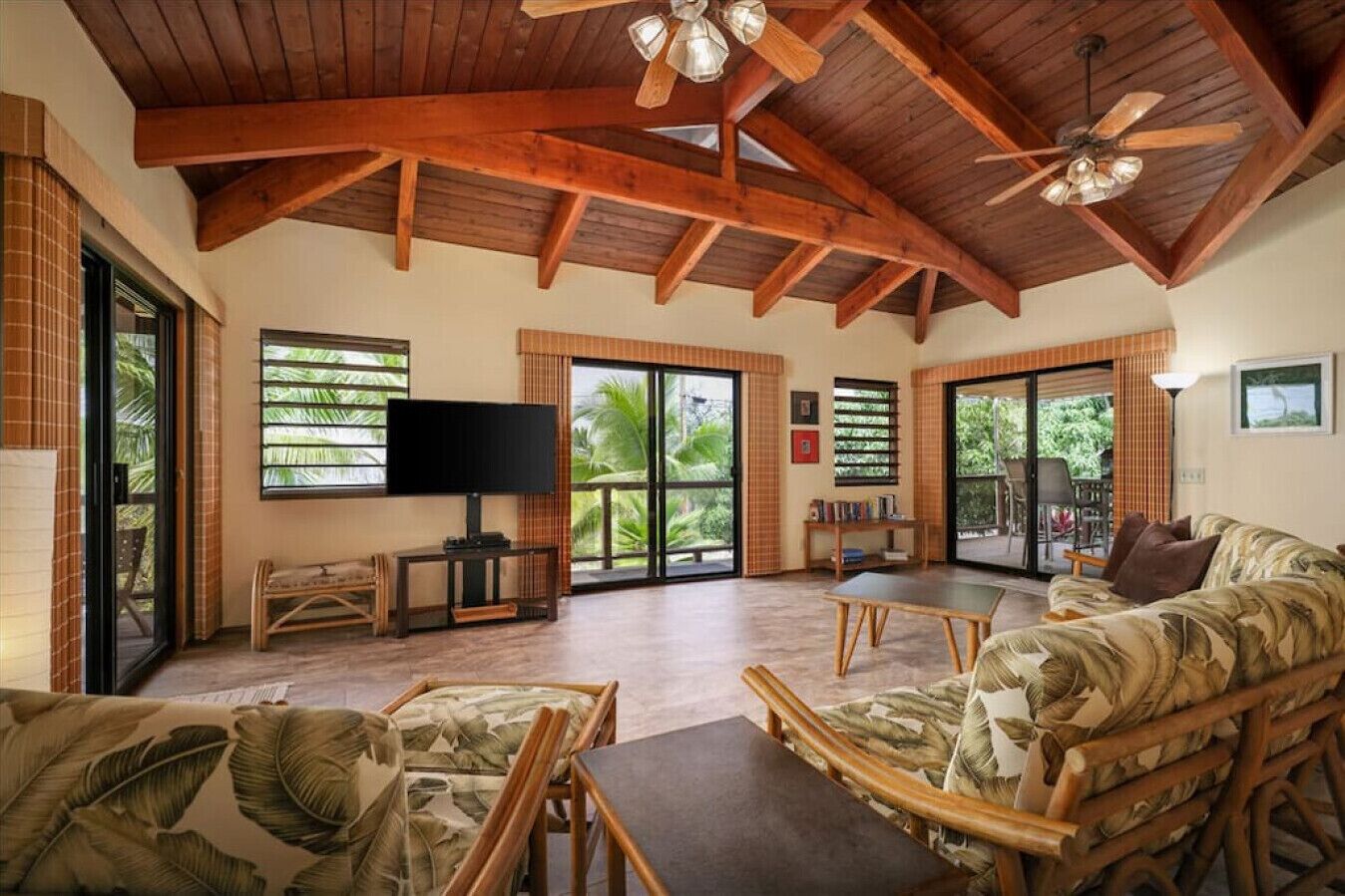 A spacious living area with a high ceiling, wooden beams, a ceiling fan, a flat-screen TV, a coffee table, a sofa with a leaf pattern, and large sliding glass doors leading to a balcony.