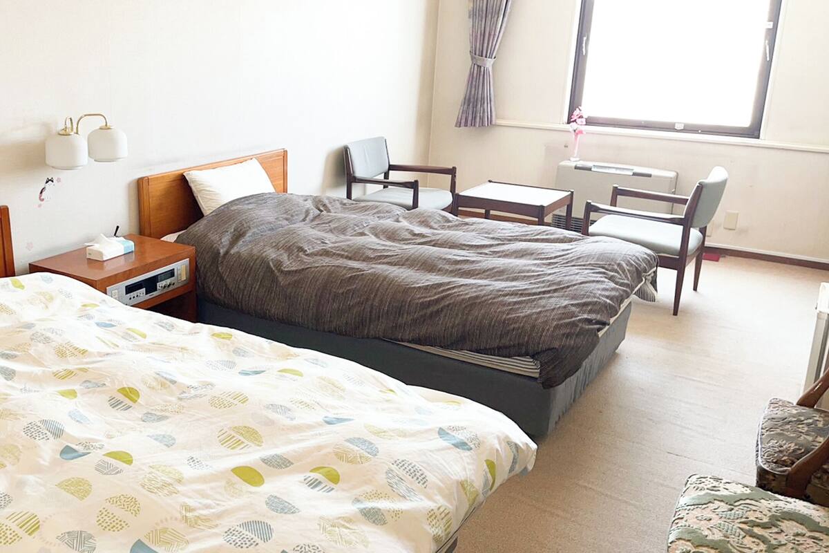 Habitación de hotel con dos camas, una mesita de noche, una silla, una pequeña mesa y una ventana con cortinas.
