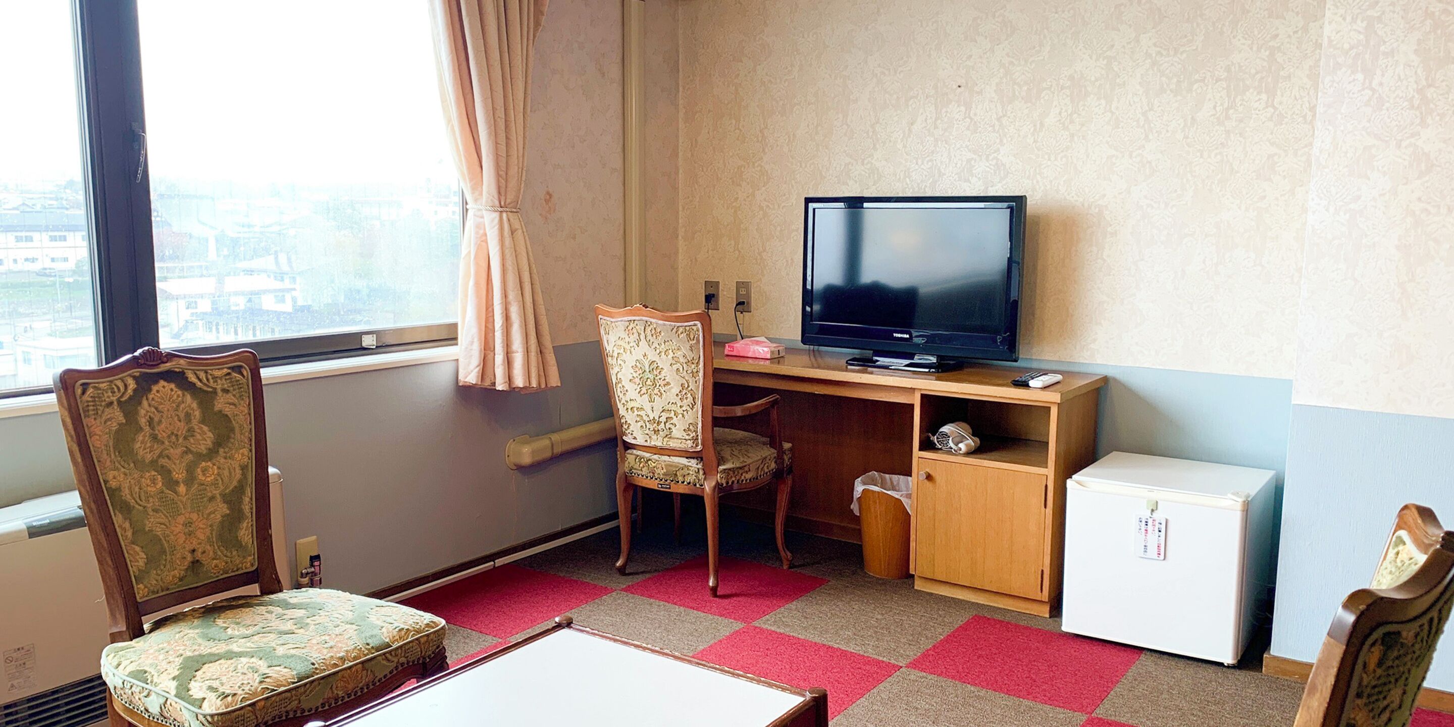 A hotel room with a wooden desk, a television, a small refrigerator, a chair, and a view of the city through a large window.