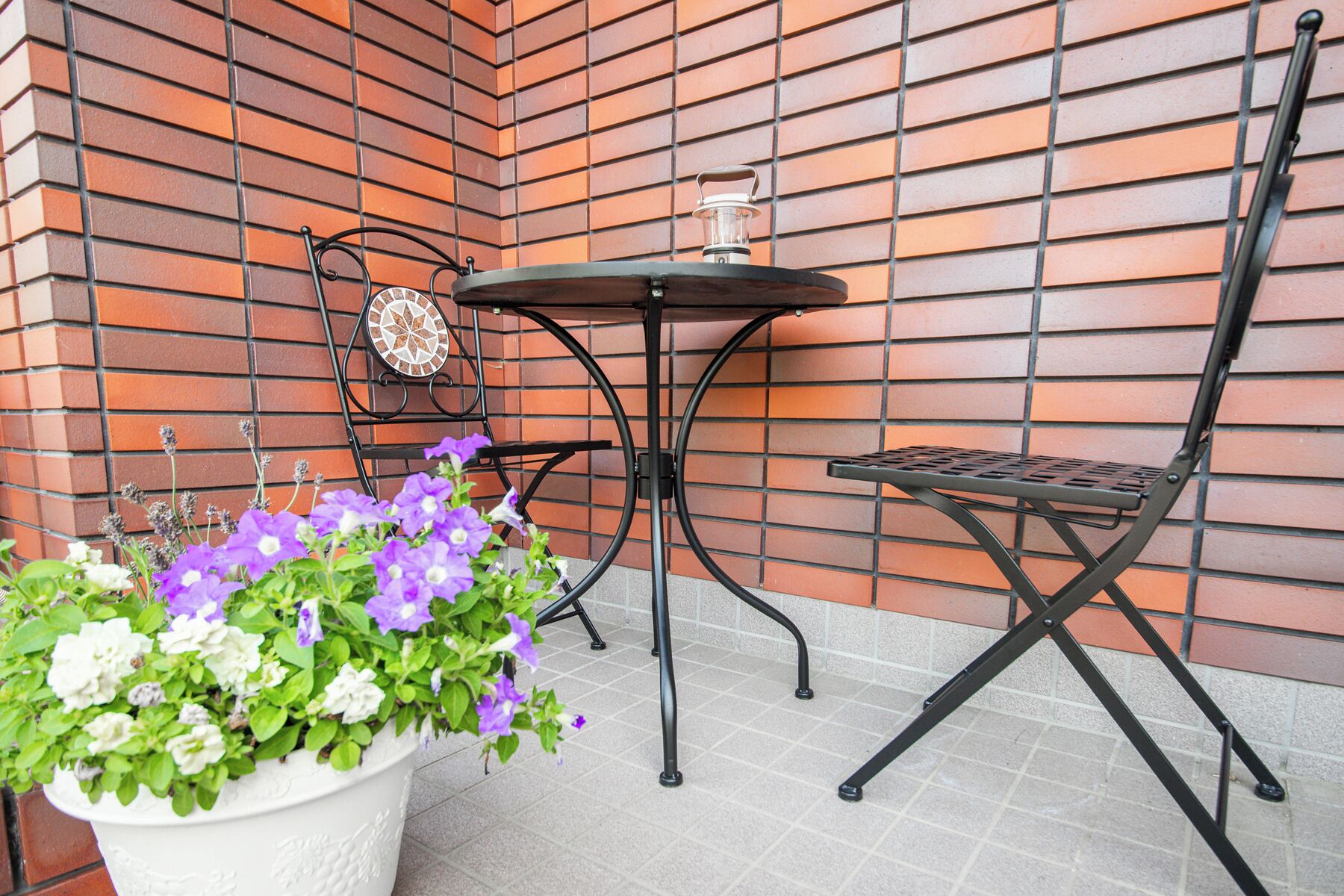 A small outdoor seating area with a metal table and chairs, a potted plant with flowers, and a decorative tile wall.