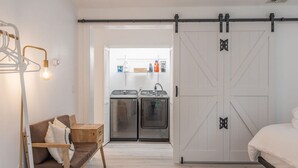 Une chambre comprenant un lit, un banc en bois, un luminaire et une porte coulissante de style rustique donnant sur un espace buanderie équipé de deux machines à laver.