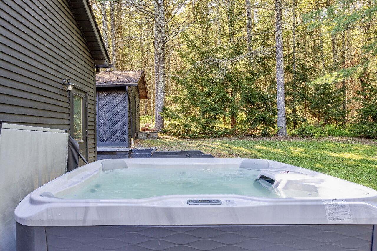 A hot tub in a backyard with a wooden house and a forest in the background.