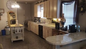 A kitchen with wooden cabinets, a coffee station, a dishwasher, and a large window with blinds.