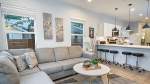 Una sala de estar moderna con un sofá gris, una mesa de centro redonda y una zona de cocina con mostrador tipo bar y taburetes.