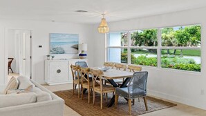 Un espace repas comprenant une table et des chaises, un meuble blanc, un tableau et une vue sur la verdure grâce à de grandes fenêtres.