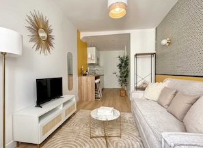 Ein Wohnzimmer mit Fernseher, Sofa und einem kleinen Couchtisch. Im Hintergrund ist eine Küche mit einer Barbillett zu sehen.