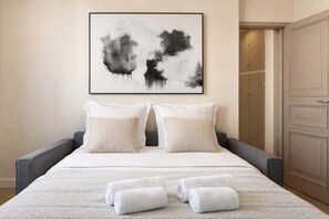 A neatly made bed with white linens and pillows, a framed abstract painting above it, and a set of towels on the bed. - Elegant property - Eiffel Tower & Invalides (Paris)
