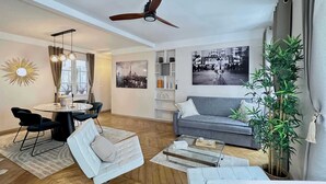 Un salon moderne comprenant un canapé, une table basse en verre, un coin repas avec des chaises, un ventilateur de plafond et une grande fenêtre.