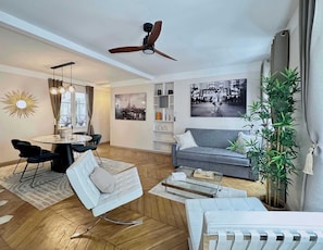 Un salon moderne comprenant un canapé, une table basse en verre, un coin repas avec des chaises, un ventilateur de plafond et une grande fenêtre.