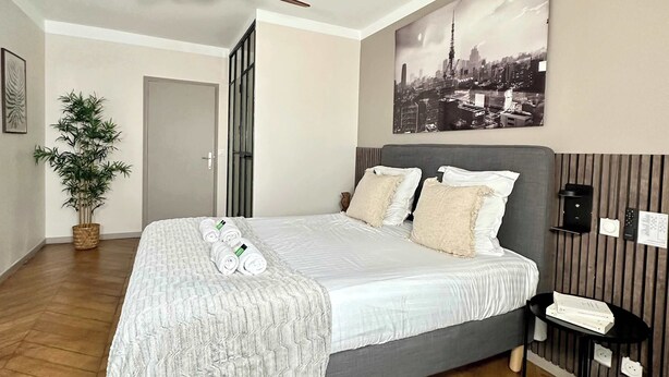 Une chambre d’hôtel avec un lit, un ventilateur de plafond, un tableau représentant un paysage urbain, une plante en pot et un plancher en bois.