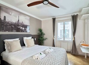 Habitación de hotel con cama, ventilador de techo, ventana con cortinas, una planta y un cuadro de paisaje urbano en la pared.