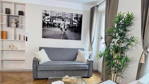 Un salon moderne avec un canapé gris, une table basse en verre, un fauteuil blanc, une grande plante en pot et une photographie en noir et blanc accrochée au mur.
