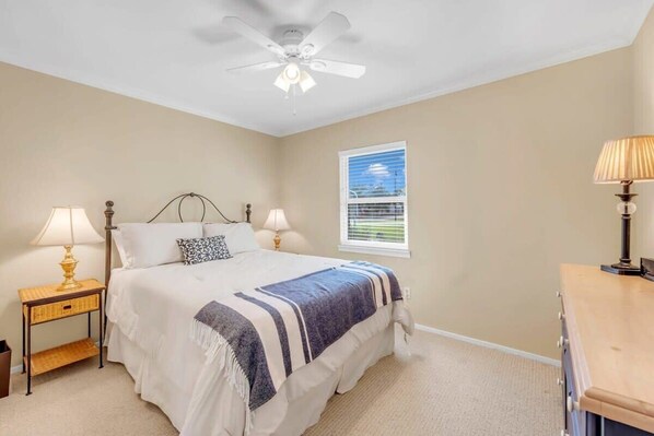 Une chambre à coucher comprenant un lit, deux tables de chevet avec des lampes, un ventilateur de plafond, une fenêtre avec des stores et une commode.