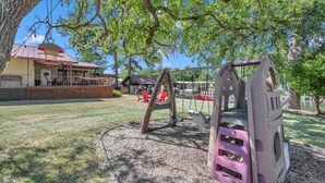 A playground with a swing set and a climbing structure, surrounded by trees and a building with a red roof.