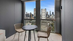 Un balcón con dos sillas y una mesa, ventanas amplias que dan a vistas de la ciudad, y una pared con textura.