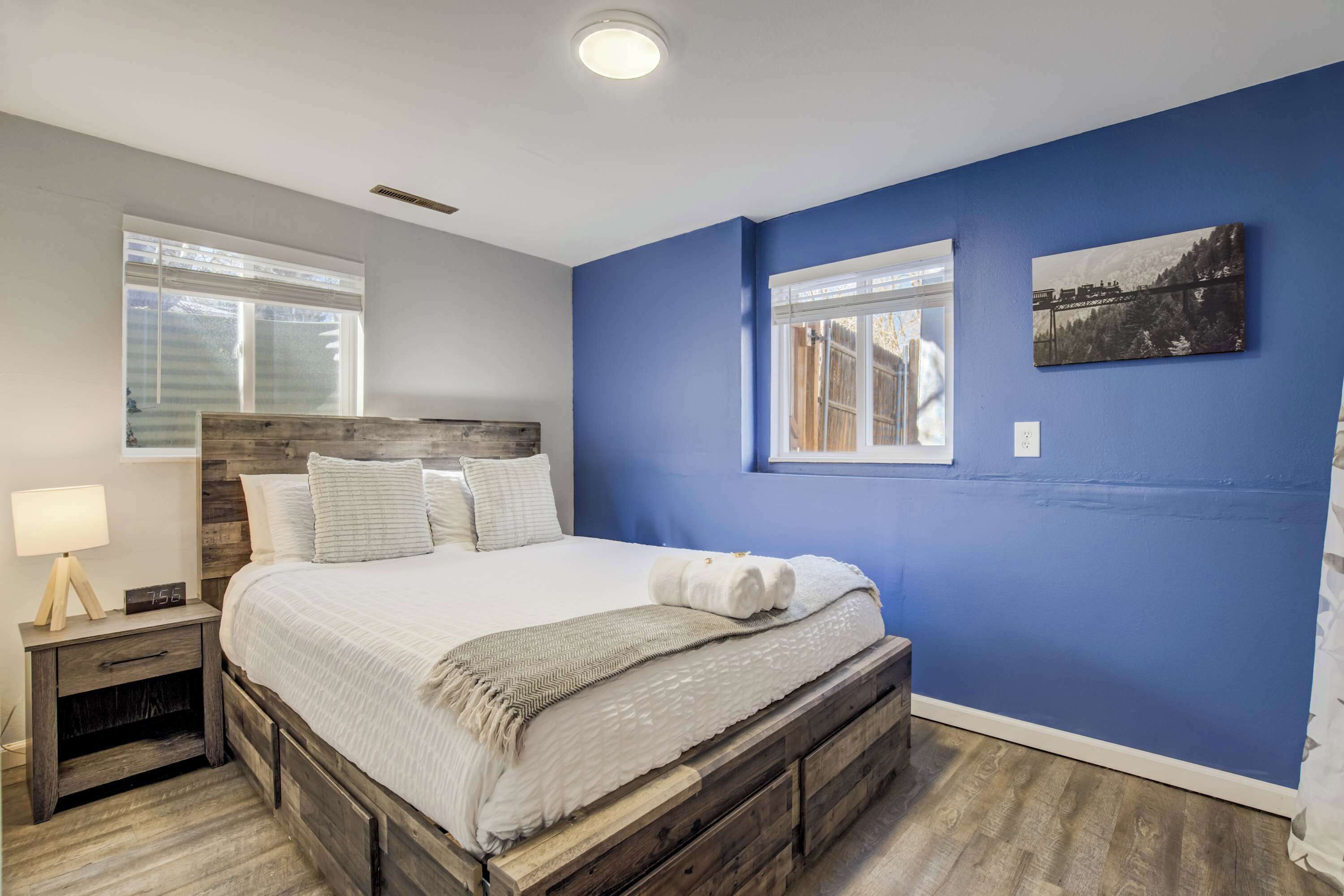 Une chambre à coucher avec un cadre de lit en bois, du linge de lit blanc, une table de chevet avec une lampe et un mur bleu orné d’un tableau.