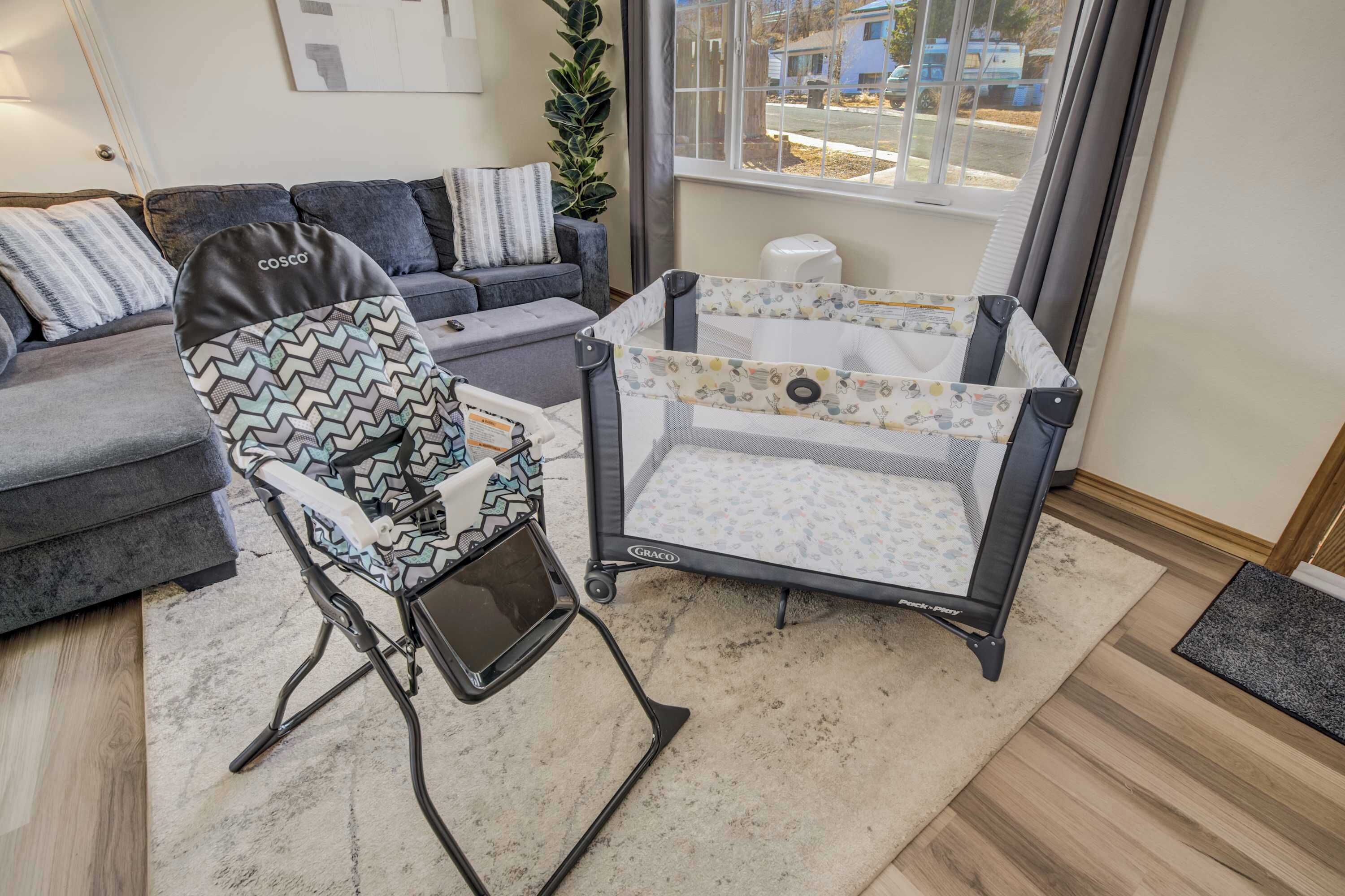 Uma muda de fraldas com berço e cadeirinha de refeição, um sofá cinzento e uma janela com vista para casas.