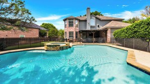 Une piscine avec un jacuzzi, entourée d’une cour clôturée, et une maison avec une grande cheminée en arrière-plan.