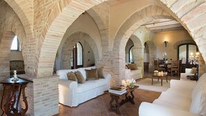 Een ruime woonkamer met witte banken, een bijzettafeltje en een open haard. De kamer heeft bakstenen muren en gewelfde plafonds.