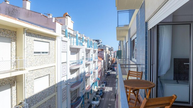 Ein Balkon mit Holztisch und -stühlen, der auf eine Straße mit parkenden Autos und Häusern mit Balkonen blickt.