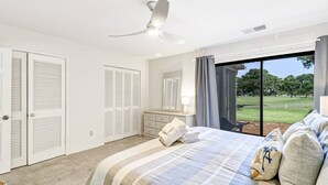 A bedroom with a bed, a nightstand, a mirror, a ceiling fan, and a view of a grassy area through a large window.