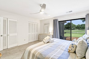 Un dormitorio con cama, mesita de noche, espejo, ventilador de techo y vistas a un área verde a través de un ventanal grande.