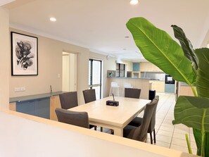 A modern dining area with a table and chairs, a kitchen in the background, and a large green plant in the foreground.