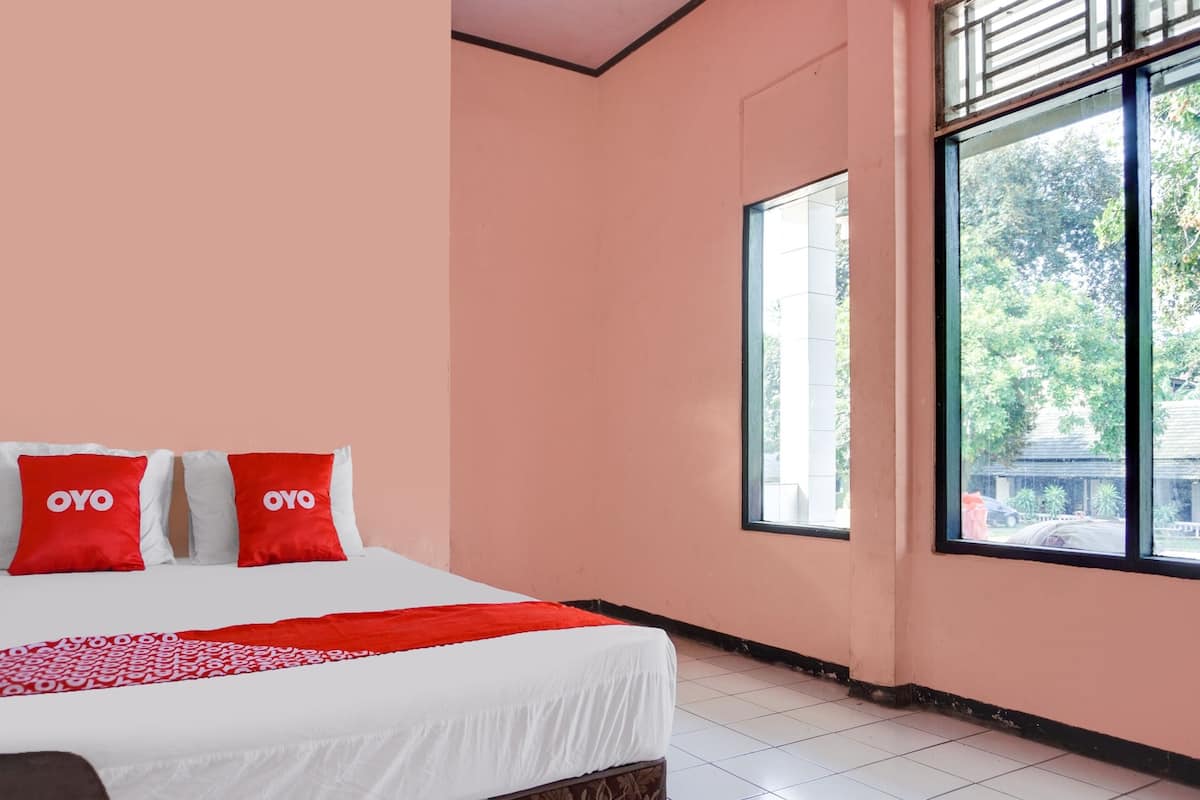Une chambre à coucher avec un lit, deux oreillers rouges ornés du texte « OYO », une couverture rouge et blanche à motifs, et une grande fenêtre donnant sur des arbres et des bâtiments.