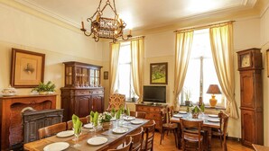 Ein Esszimmer mit einem langen Holztisch, gedeckt für eine Mahlzeit, umgeben von Stühlen. Eine Pendelleuchte hängt von der Decke, und es gibt einen Kamin mit Kaminfeuer.
