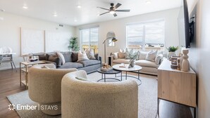 Un vaste salon avec un canapé gris, des fauteuils beiges et une table basse. On y trouve un ventilateur de plafond, un téléviseur à écran plat fixé au mur et de grandes fenêtres offrant une vue sur le paysage désertique.