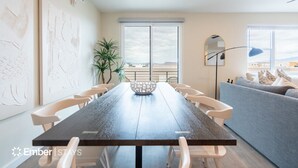 Un espace de repas moderne avec une longue table, des chaises blanches et un grand vase décoratif au centre. Une grande fenêtre offre une vue sur le paysage, et on aperçoit une plante en pot et un canapé bleu à l’arrière-plan.