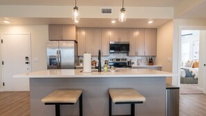 Une cuisine moderne dotée d’un grand îlot central, d’appareils électroménagers en acier inoxydable et de luminaires suspendus.
