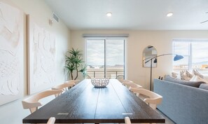 A modern dining area with a long table, white chairs, and a central decorative bowl. There is a large window with a view of the landscape, a potted plant, and a blue sofa in the background.